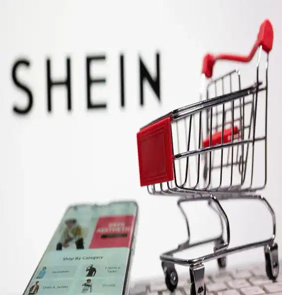 Imagem mostra um celular em primeiro plano, um carrinho de compras atrás e um letreiro escrito Shein. Foto: Reuters/Ilustra/Arquivo